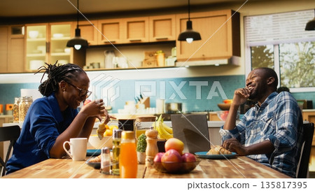 African american couple enjoying coffee and breakfast together at kitchen table, laughing and chatting in a cozy apartment. Feeling relaxed in the morning, positive energy indoors. 135817395