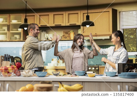 Pleased family of three sharing a high five and cheering in the kitchen, finishing homemade pizza for pizza night. Parents and pre-teen girl celebrating their teamwork with laughter. Pleased family of three sharing a high five and cheering in the kitchen, finishing homemade pizza for pizza night. Parents and pre-teen girl celebrating their teamwork with laughter. 135817400