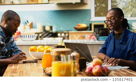 Mature boyfriend and girlfriend sitting together at kitchen table with paperwork and laptop. Tracking expenses, calculating bills and managing household money, responsible fiscal planning. 135817401