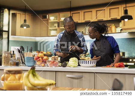 Black boyfriend and girlfriend preparing a meal with online food recipe on tablet, cutting ingredients and making a vegetarian salad in the kitchen, healthy lifestyle and culinary bonding. Black boyfriend and girlfriend preparing a meal with online food recipe on tablet, cutting ingredients and making a vegetarian salad in the kitchen, healthy lifestyle and culinary bonding. 135817490
