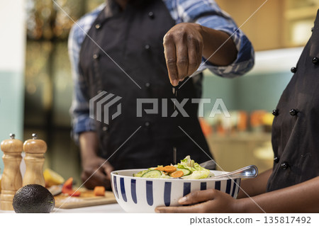 African american man seasoning a salad with salt in a bowl, preparing a healthy lunch at home with organic vegetables. Young vegetarian couple eating fresh produce, wellness and balance. African american man seasoning a salad with salt in a bowl, preparing a healthy lunch at home with organic vegetables. Young vegetarian couple eating fresh produce, wellness and balance. 135817492