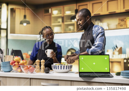 Green screen on laptop next to black couple preparing food in the kitchen, making a fresh organic salad with bio ingredients and veggies. Positive joyful man and woman with aprons. Green screen on laptop next to black couple preparing food in the kitchen, making a fresh organic salad with bio ingredients and veggies. Positive joyful man and woman with aprons. 135817498