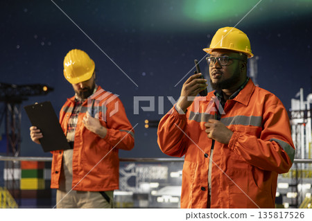 Radio operator working on drilling derrick on ocean provides operational updates with walkie talkie. African american man on offshore platform maintains radio contact at night under aurora borealis Radio operator working on drilling derrick on ocean provides operational updates with walkie talkie. African american man on offshore platform maintains radio contact at night under aurora borealis 135817526