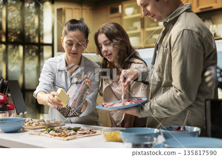 Parents and pre-teen daughter sprinkling grated cheese and mozzarella on fresh pizza dough, laughing in the kitchen together in a cute family moment filled with joy and culinary creativity. Parents and pre-teen daughter sprinkling grated cheese and mozzarella on fresh pizza dough, laughing in the kitchen together in a cute family moment filled with joy and culinary creativity. 135817590