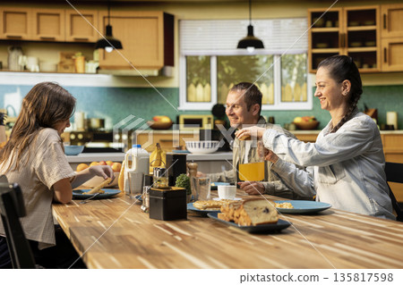 Mom and dad gathered at the breakfast table with daughter to eat waffles and fresh homemade food. Kitchen scene shows morning connection, family bonding to share homemade meal together. Mom and dad gathered at the breakfast table with daughter to eat waffles and fresh homemade food. Kitchen scene shows morning connection, family bonding to share homemade meal together. 135817598