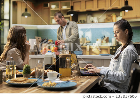 Distracted mom ignoring her pre-teen girl at breakfast and focusing on her task, typing information on laptop and looking preoccupied. Absent busy mother with a career and tween girl. 135817600