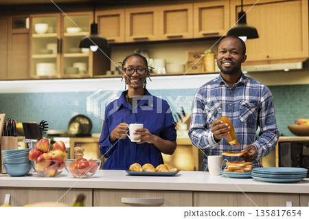 Portrait of African American lovers standing close in the kitchen, enjoying morning coffee and handmade pastry with honey. Joyful couple smiling before serving a delicious homemade breakfast. Portrait of African American lovers standing close in the kitchen, enjoying morning coffee and handmade pastry with honey. Joyful couple smiling before serving a delicious homemade breakfast. 135817654