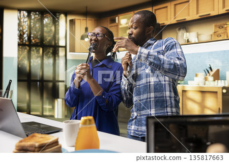 Goofy cheerful people singing together with tools as microphones in the kitchen, black couple showing love and playful energy, fooling around and having fun with music. Positive relations. 135817663