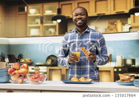 Portrait of african american man holding a glass of orange juice and a croissant, smiling in the kitchen. Morning routine expresses calmness and happiness in comfortable homey atmosphere. Portrait of african american man holding a glass of orange juice and a croissant, smiling in the kitchen. Morning routine expresses calmness and happiness in comfortable homey atmosphere. 135817664