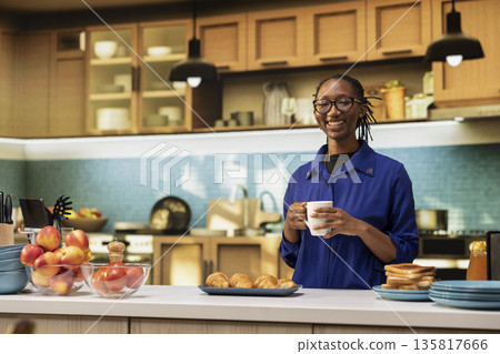 Portrait of smiling black woman serving fresh coffee in a mug before preparing toast and pastry for breakfast. Enjoying calm morning at home, surrounded by kitchenware and healthy food. 135817666
