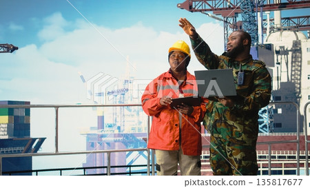 Military personnel stationed on offshore rig analyzing tactical data on laptop. Soldier provides protection to drilling barge, reviewing mission dashboard on notebook, 135817677