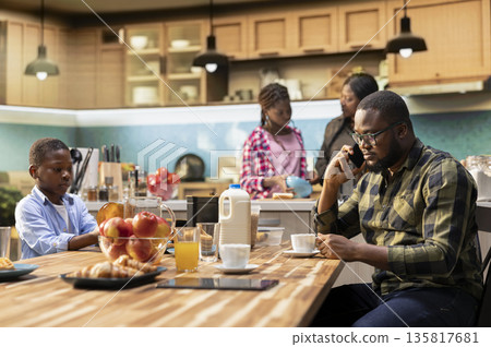 Black young dad on a phone call for work during breakfast meal together, interrupting family bonding time and morning routine to answer important calls. Father neglecting his children. 135817681