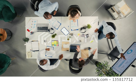 Top down view of business colleagues share insight on finances and payments during meeting. Staff review documents, diagrams and reports to align targets and planning strategies. Top down view of business colleagues share insight on finances and payments during meeting. Staff review documents, diagrams and reports to align targets and planning strategies. 135817712