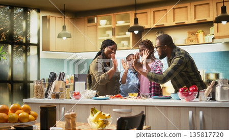 Portrait of black family clapping hands and cheering after finishing homemade pizza in the kitchen. High five and teamwork with laughter shows joy of family cooking and bonding. Camera A. Portrait of black family clapping hands and cheering after finishing homemade pizza in the kitchen. High five and teamwork with laughter shows joy of family cooking and bonding. Camera A. 135817720