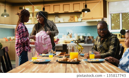 African American mother packing lunch box for her two school kids, preparing their backpack with essentials and snacks for the day. Enjoying rich breakfast with family at home. Camera A. 135817743