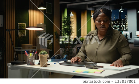 Black businesswoman closing her laptop at the office workstation, finishing work after hours at midnight and going home. Leaving the workstation building after doing overtime. Camera A. Black businesswoman closing her laptop at the office workstation, finishing work after hours at midnight and going home. Leaving the workstation building after doing overtime. Camera A. 135817801