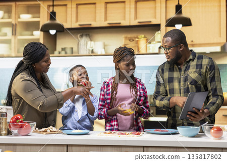 Black adorable parents and kids following online cooking recipe for pizza day, enjoying fun weekend activity making homemade pizza together in the kitchen. Happiness and teamwork for bonding. Black adorable parents and kids following online cooking recipe for pizza day, enjoying fun weekend activity making homemade pizza together in the kitchen. Happiness and teamwork for bonding. 135817802