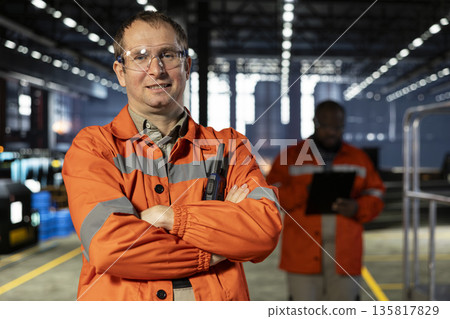 Industrial personnel with safety vest and safety glasses in fabrication workshop, overseeing machinery as sparks rise from metal welding. Manufacturing process and workforce labor. Industrial personnel with safety vest and safety glasses in fabrication workshop, overseeing machinery as sparks rise from metal welding. Manufacturing process and workforce labor. 135817829