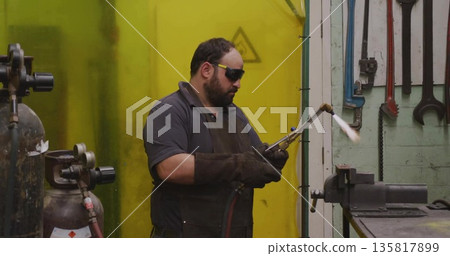 Operating metalworker in leather apron heating vise in workshop, with blowtorch and gas cylinders 135817899