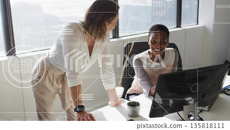 Collaborating two mid adult women wearing blouses leaning over PC at office desk, with coffee mug 135818111