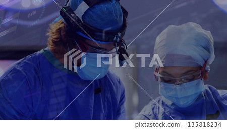 Operating surgeons in blue gowns performing surgery in operating room, with headlight instruments Operating surgeons in blue gowns performing surgery in operating room, with headlight instruments 135818234