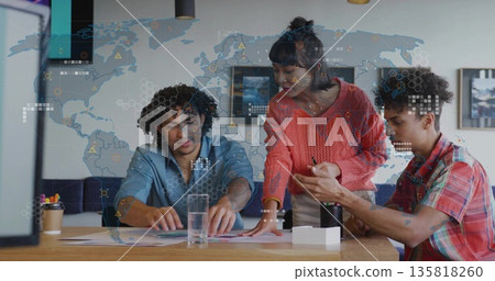 Collaborating team analyzing charts at office desk with water glass, tumbler, supplies, map overlay 135818260