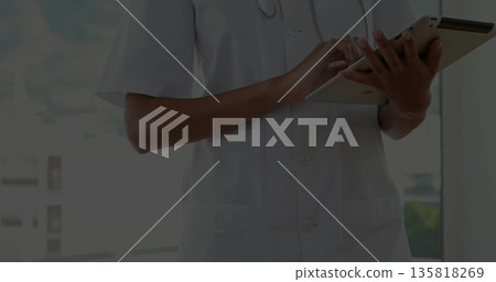 Typing healthcare professional wearing white coat and stethoscope in clinic by window, with tablet Typing healthcare professional wearing white coat and stethoscope in clinic by window, with tablet 135818269