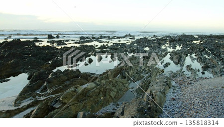 Revealing jagged rock formations and tide pools reflecting sky at low tide, with pebbles and shells 135818314