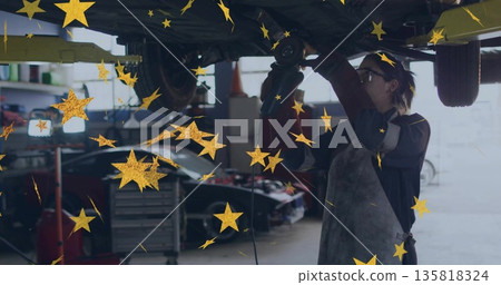 Reaching technician wearing apron goggles operating drill under car on lift in bay with tool chest 135818324