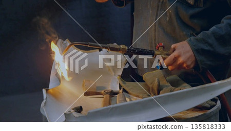 Shaping metal panel metalworker wearing jacket using blowtorch on drum in shop, with hammers mallet 135818333