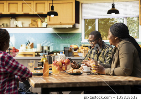 Black distant father paying attention to smartphone and ignoring his family during breakfast meal. Absorbed preoccupied working dad distracted by mobile phone and apps, neglecting children. 135818532