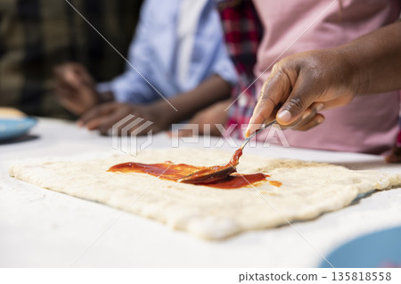 Close up of african american parents and kids using tomato sauce on the dough, preparing a homemade pizza with fresh ingredients before baking. Family fun and bonding time at home. Close up of african american parents and kids using tomato sauce on the dough, preparing a homemade pizza with fresh ingredients before baking. Family fun and bonding time at home. 135818558