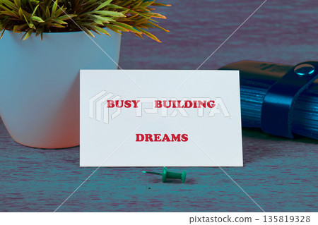 White card displaying Busy Building Dreams in red stands on a desk. A green pushpin sits below White card displaying Busy Building Dreams in red stands on a desk. A green pushpin sits below 135819328