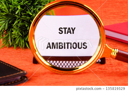 A magnifying glass highlights the words STAY AMBITIOUS on a white paper. It sits on a bright orange table alongside a green plant and red notebook indoors during the day 135819329