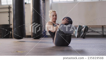 Fitness trainee performing seated Russian twisting in gym, holding medicine ball near punching bags Fitness trainee performing seated Russian twisting in gym, holding medicine ball near punching bags 135819588