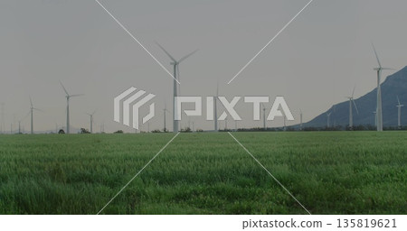 Towering white wind turbine rotating three blade rotor over rural grass field, with mountain range Towering white wind turbine rotating three blade rotor over rural grass field, with mountain range 135819621