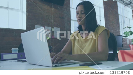 Typing mature Asian woman using silver laptop at modern open-plan office desk, with coffee cup Typing mature Asian woman using silver laptop at modern open-plan office desk, with coffee cup 135819708