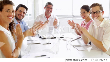 Applauding group of six professionals seated around conference table in office with notebooks, pens Applauding group of six professionals seated around conference table in office with notebooks, pens 135819744