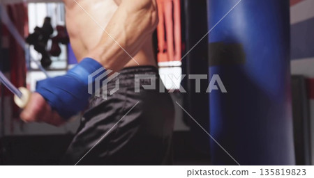 Skipping shirtless man jumping rope at boxing gym near punching bag, wearing blue hand wraps Skipping shirtless man jumping rope at boxing gym near punching bag, wearing blue hand wraps 135819823