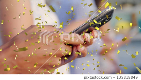 Gripping brown-skinned hand holding smartphone in warm interior, with floating golden confetti Gripping brown-skinned hand holding smartphone in warm interior, with floating golden confetti 135819849