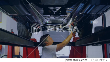 Inspecting mechanic in gray shirt operating wrench under vehicle at garage with red lift supports Inspecting mechanic in gray shirt operating wrench under vehicle at garage with red lift supports 135819937