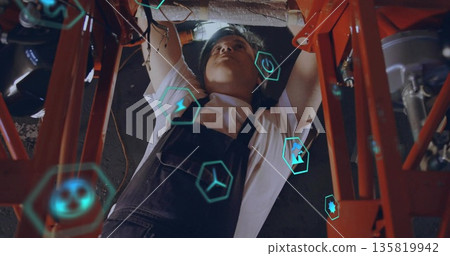 Technician in overalls adjusting pipes in workshop under orange beams and ducts, with digital icons 135819942