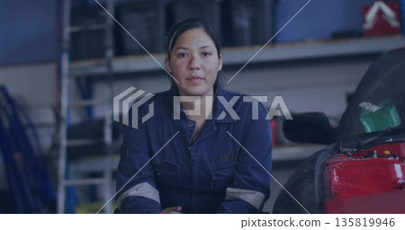 Kneeling female mechanic wearing navy coveralls inspecting red car engine in shop, with shelves 135819946