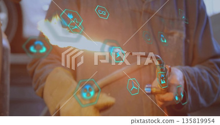 Technician wearing gloves and jacket holding torch emitting flame in metal workshop, with AR icons Technician wearing gloves and jacket holding torch emitting flame in metal workshop, with AR icons 135819954