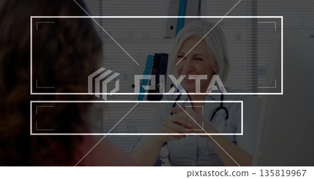 Smiling physician wearing gray tunic consulting patient at clinic desk, with stethoscope, binders 135819967