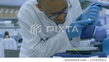 Leaning scientist in lab coat dispensing liquid into 96-well plate at lab bench, with pipette Leaning scientist in lab coat dispensing liquid into 96-well plate at lab bench, with pipette 135820022
