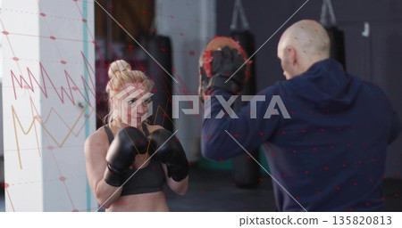 Training boxer wearing sports bra hitting focus mitts at boxing gym, with gloves and punching bags 135820813