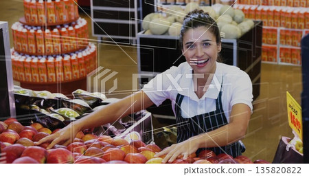 Organizing clerk in green-striped apron leaning over red apples at market produce, with juice trays Organizing clerk in green-striped apron leaning over red apples at market produce, with juice trays 135820822
