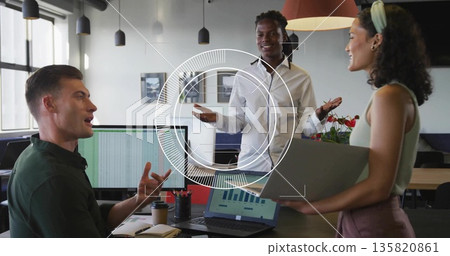 Three coworkers in formal wear discussing insights at desk in office, with laptop monitors 135820861