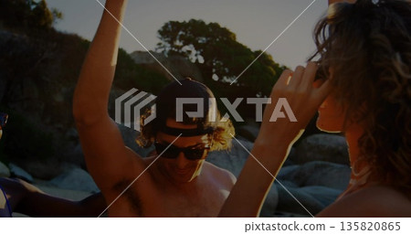 Posing man wearing swim trunks, baseball cap and sunglasses raising arm in rocky cove with friends Posing man wearing swim trunks, baseball cap and sunglasses raising arm in rocky cove with friends 135820865